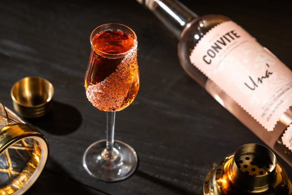 A rose colored cocktail with a bottle of Una'